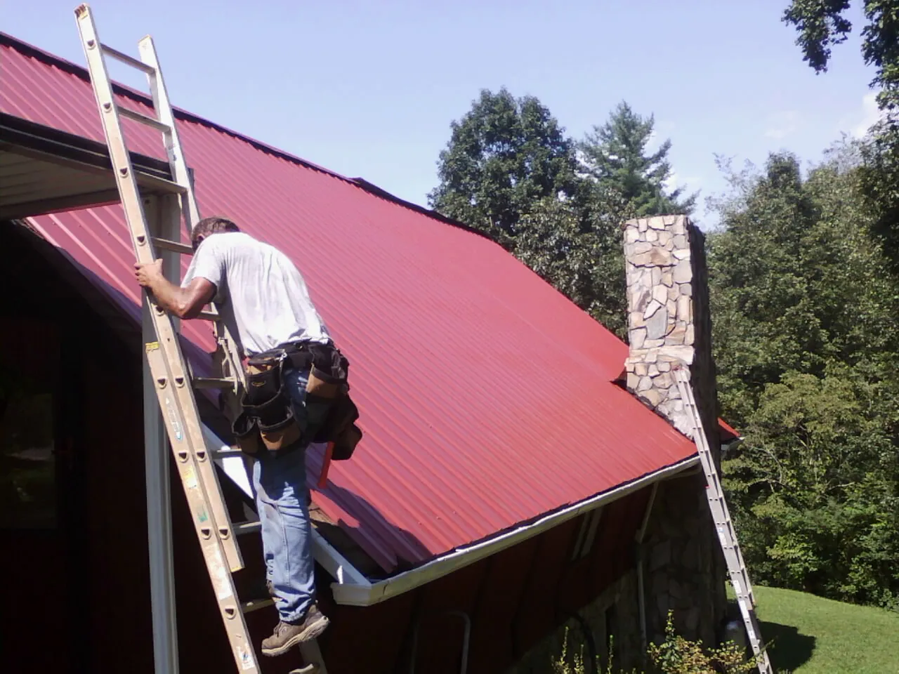 Roofer on a ladder beside a red metal roof and stone chimney — Soffit & Fascia Repair by Roofing Brothers in Mount Olive